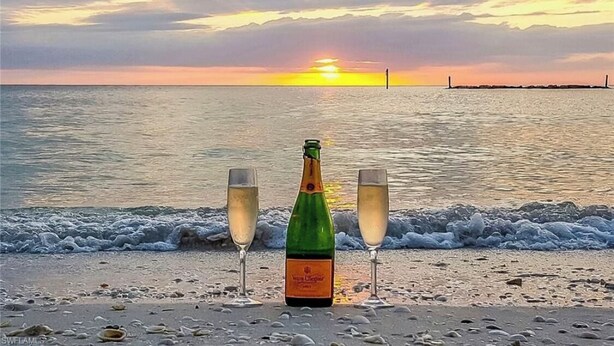 En flaska champagne och två glas på en strand, med havet och solnedgången i bakgrunden.