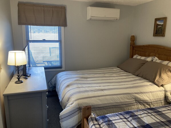 Ein Schlafzimmer mit Bett, Nachttisch, Lampe, Fenster mit Jalousien und einer Klimaanlage.
