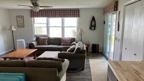 A living room with a sectional sofa, a wooden coffee table, and a ceiling fan. There is a window with a view of the outdoors. - Spring Summer specials on cozy newly renovated cottage with AC right on the lake (Honeoye)