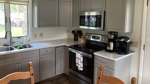 A kitchen with grey cabinets, a stainless steel microwave, a stove, and a coffee maker. - Spring Summer specials on cozy newly renovated cottage with AC right on the lake (Honeoye)