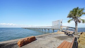 A concrete pier extending into calm waters, a wooden bench, a palm tree, and a clear sky. - Awesome Bay View Home with Two Living Areas!  Pets welcome! (Galveston)