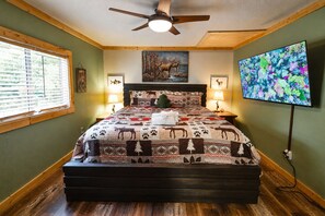 A bedroom with a large bed, a ceiling fan, a painting, a television, and a window with blinds. - Walkable Pigeon Forge Escape + Hot Tub (Pigeon Forge)