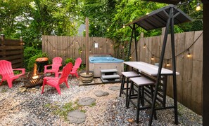An outdoor patio with a fire pit, red chairs, a hot tub, and a wooden fence.