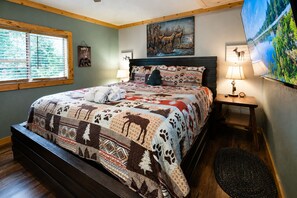 A bedroom with a large bed, a nightstand, a lamp, a painting, and a window with blinds.