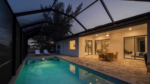 A modern outdoor pool area with a glass-enclosed patio, lounge chairs, and a dining area with a table and chairs. - Beautiful modern pool home; Furnished lanai, gas grill; Free Wifi; Nearby park (Cape Coral)