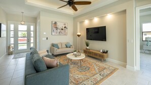 A spacious living room with a ceiling fan, a large TV mounted on the wall, a sofa, a coffee table, and a patterned rug. - Beautiful modern pool home; Furnished lanai, gas grill; Free Wifi; Nearby park (Cape Coral)