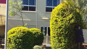 A building with a red roof, a grey facade, and a central entrance. There are two well-maintained bushes in front of the entrance, and a palm tree to the right.