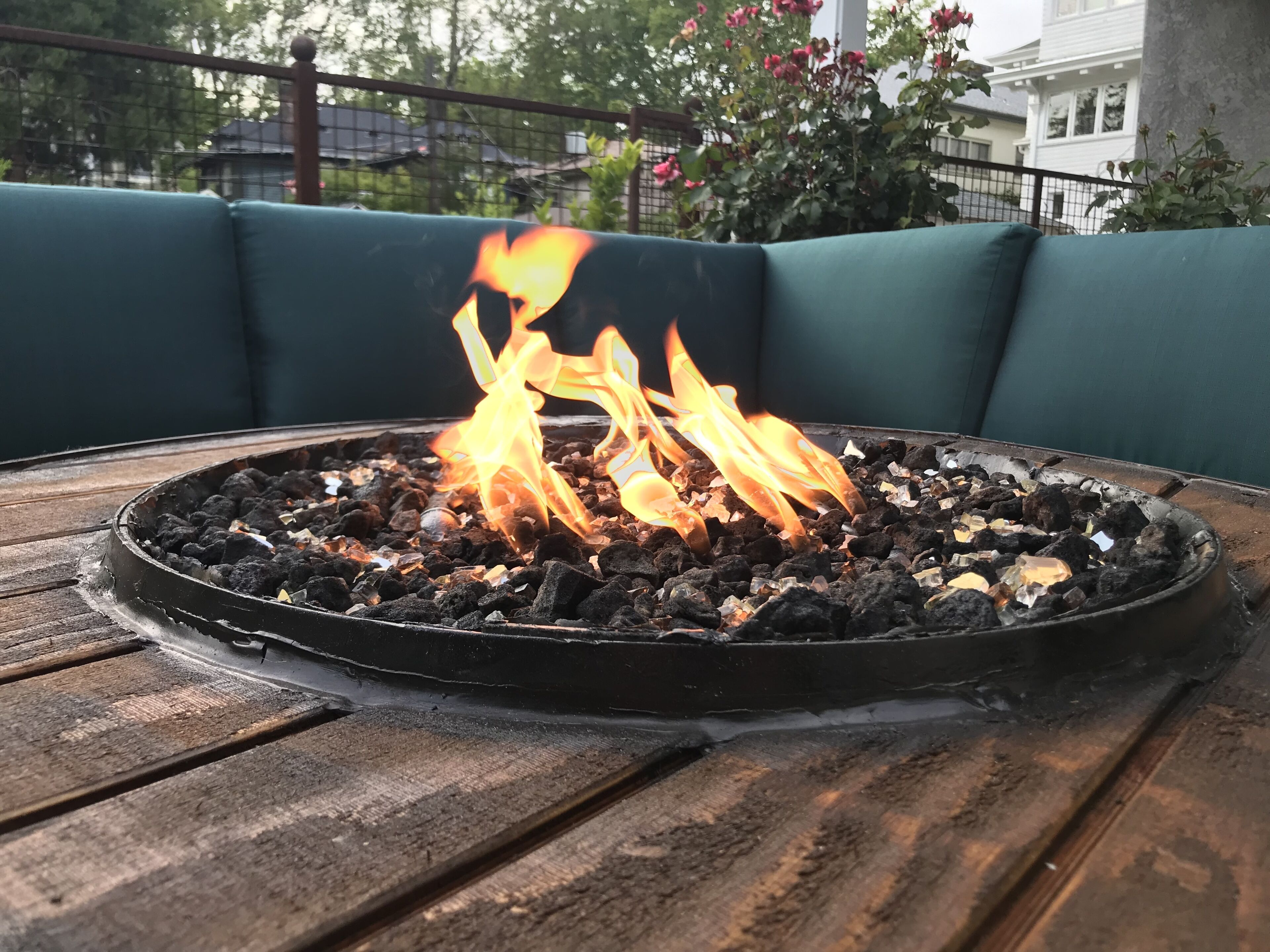 Un pozo de fuego circular de carbón con llamas sobre una mesa de madera, rodeado de asientos verdes para exteriores.