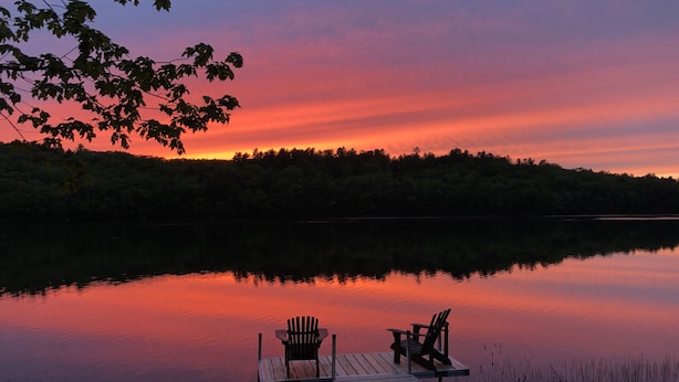 A wooden dock extending into a calm lake with two chairs facing the water, surrounded by trees and a sunset sky.