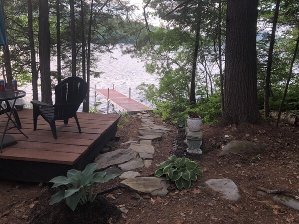 A wooden deck with chairs, a stone pathway, and a view of a lake surrounded by trees.
