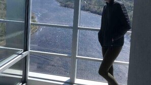 A person standing by a large window overlooking a river and surrounding landscape.