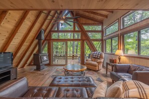 Ein gemütliches Blockhaus-Interieur mit Holzbalkendecke, großen Fenstern, Kamin, Ledermöbeln und einem Fernseher.