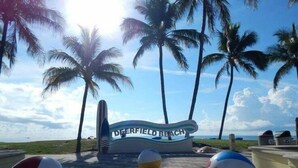 Skilt ved stranden med palmer og strandbolde i forgrunden.