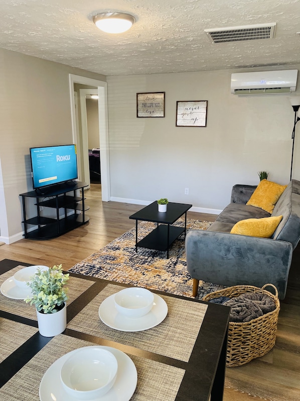A living room with a flat-screen TV, a gray sofa with a yellow pillow, a black coffee table, and a patterned rug. - Secret Hideaway (Rossville)