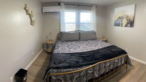 A bedroom with a bed, bedside tables, a wall-mounted air conditioner, a window with white curtains, and a painting on the wall. - Secret Hideaway (Rossville)