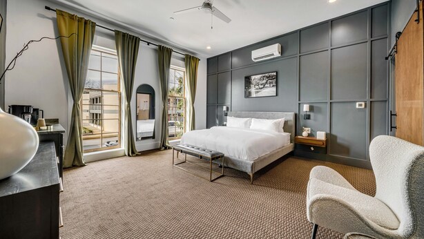 Une chambre d’hôtel moderne, dotée d’un grand lit, d’un bureau et d’une chaise. On y trouve une fenêtre avec des rideaux, un ventilateur de plafond et un appareil de climatisation fixé au mur.