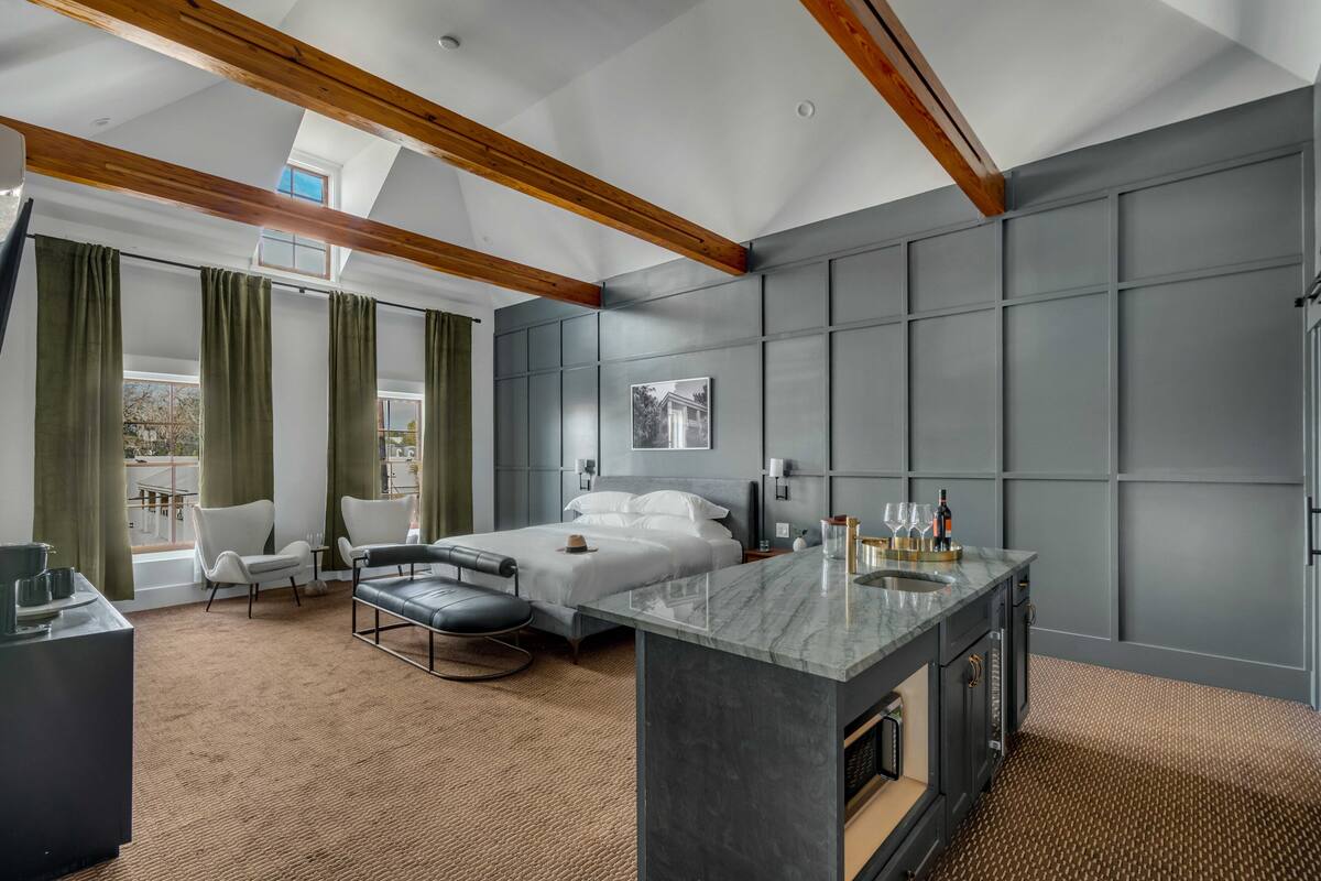 A modern hotel room with a large bed, a sitting area, a bathroom with a vanity, and a view of the outdoors.