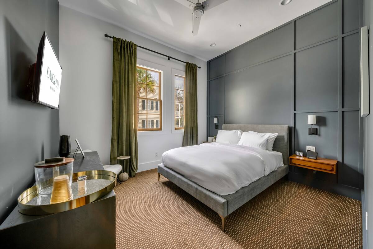 A modern hotel room with a large bed, a bedside table, a TV mounted on the wall, and a window with curtains.