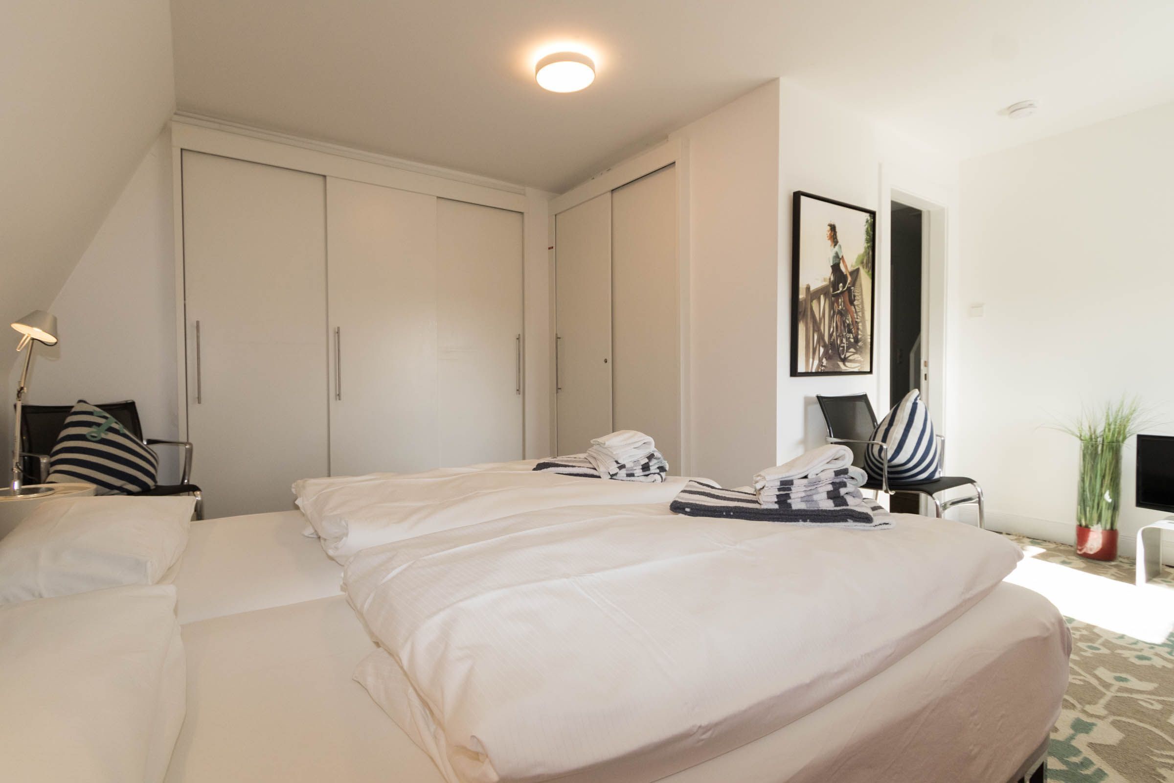 A hotel room with two beds, a desk, a chair, a lamp, a painting, and a potted plant.