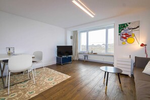 A modern living room with a flat-screen TV, a dining table with chairs, a sofa, and a large window with a view.