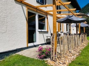 Una casa con una pérgola de madera, amplias ventanas y un jardín con flores y mantillo.