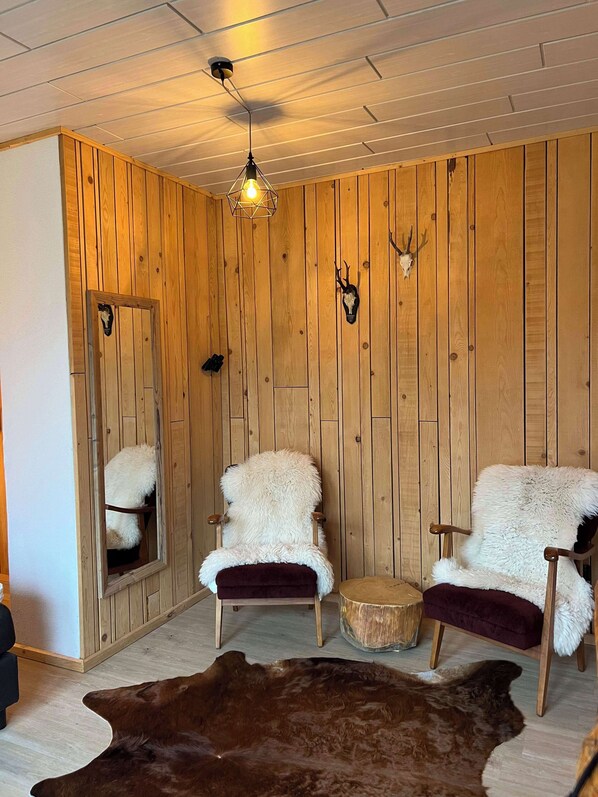 Une pièce aux murs recouverts de lambris en bois, deux fauteuils blancs à l’aspect duveteux, une table ronde en bois, un tapis en cuir marron et un luminaire suspendu au plafond.