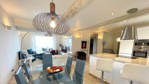 Eine moderne Küche mit einer zentralen Kochinsel, ein Essbereich mit Glastisch und Stühlen sowie ein Wohnzimmer mit Sofa und Fernseher.