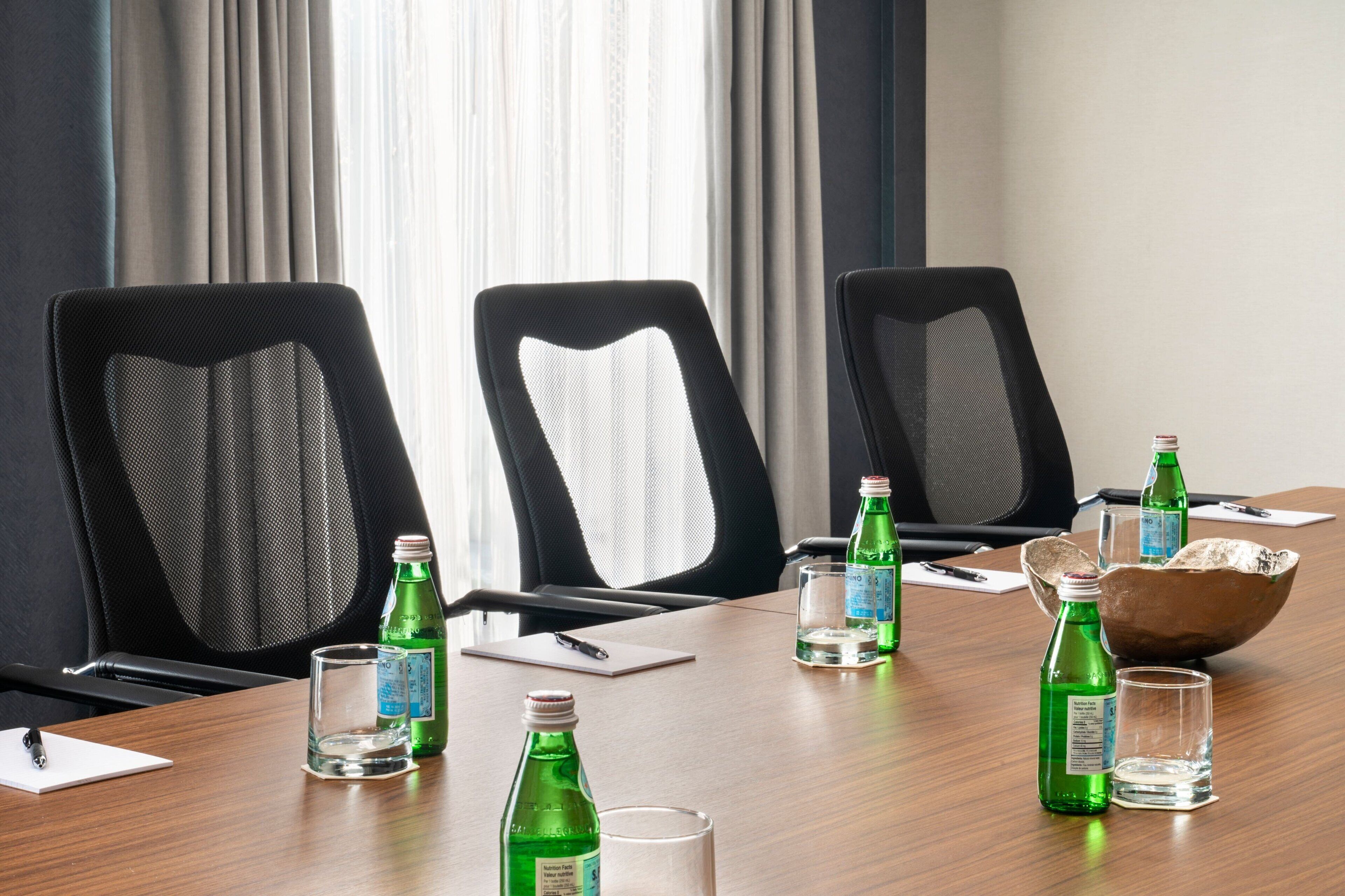 Un salón de conferencias con tres sillas de malla negra, una mesa de madera y varias botellas de agua verdes y vasos.