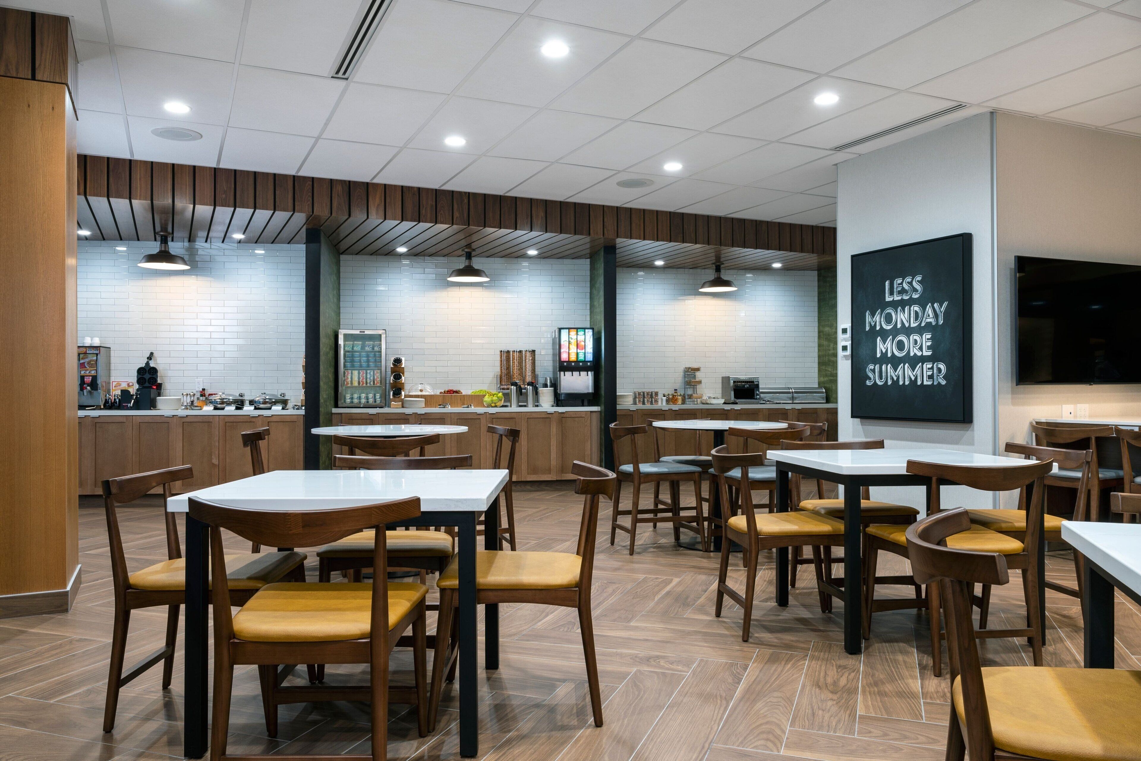 A modern restaurant interior with wooden tables and chairs, a kitchen area in the background, and a motivational sign on the wall.