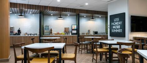 Un interior de restaurante moderno con mesas y sillas de madera, una zona de cocina al fondo y un letrero motivacional en la pared.