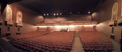 Un auditorio con hileras de asientos, un escenario y ventanas decorativas.