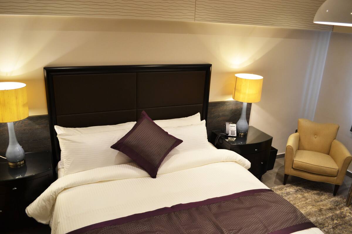A hotel room with a large bed, two bedside tables with lamps, a chair, and a carpeted floor.