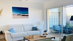 A modern living room with a white sofa, two green armchairs, a wooden coffee table, and a glass door leading to an outdoor seating area.