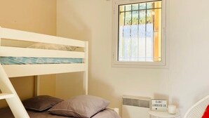 A bunk bed with a mattress and pillows, a small table with a sign, a radiator, and a window with a view of greenery.