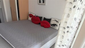 A bed with a grey bedspread, red and black pillows, and a white curtain with a leaf pattern.