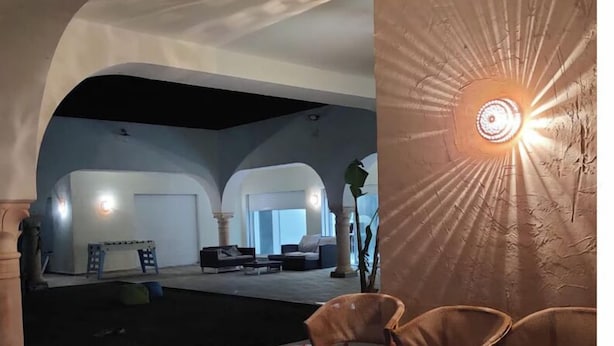 A modern interior with a central seating area, a large wall-mounted light fixture, and a view into another room with a white ceiling and furniture. - Very large villa ideal for groups of up to 15 to 18 people (Kélibia)