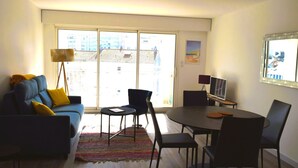 A modern living room with a blue sofa, a dining table with chairs, a floor lamp, and a large window with a city view. - 1-bed apartment, garage, direct beach access (Les Sables-d'Olonne)