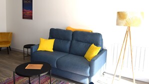 A living room with a blue sofa, yellow cushions, a small black table, a colorful rug, a yellow armchair, and a floor lamp. - 1-bed apartment, garage, direct beach access (Les Sables-d'Olonne)