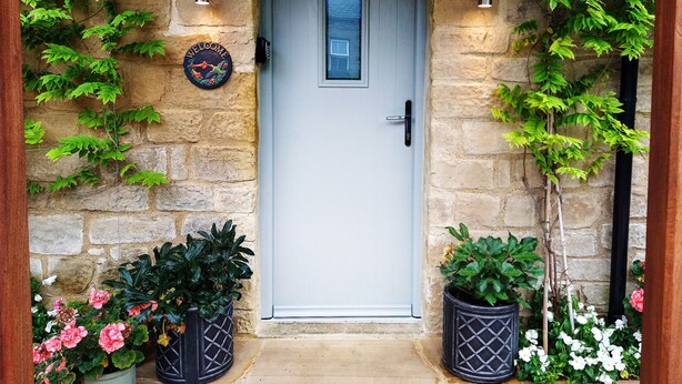 Entrada com muro de pedra, porta, plantas em vasos e placa de boas-vindas.