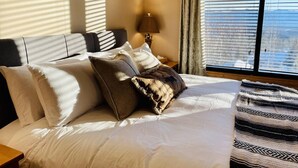 Una cama bien tendida con ropa de cama blanca y estampada, una lámpara de noche y vistas del exterior a través de una ventana con persianas.
