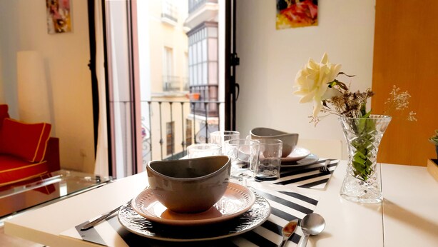 A dining table with a bowl, glasses, and cutlery, a vase with flowers, and a red sofa in a room with a view of buildings through a window. - 2 balconies! Lovely Center Apartments Alfalfa (Sevilla)