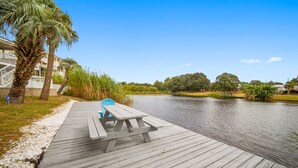 A wooden dock with a picnic table and chairs, overlooking a calm river with trees and buildings in the background. - Blue Lagoon - Lakefront Getaway Private Pool, One Block to the Beach, Sleeps 12 (Panama City Beach)