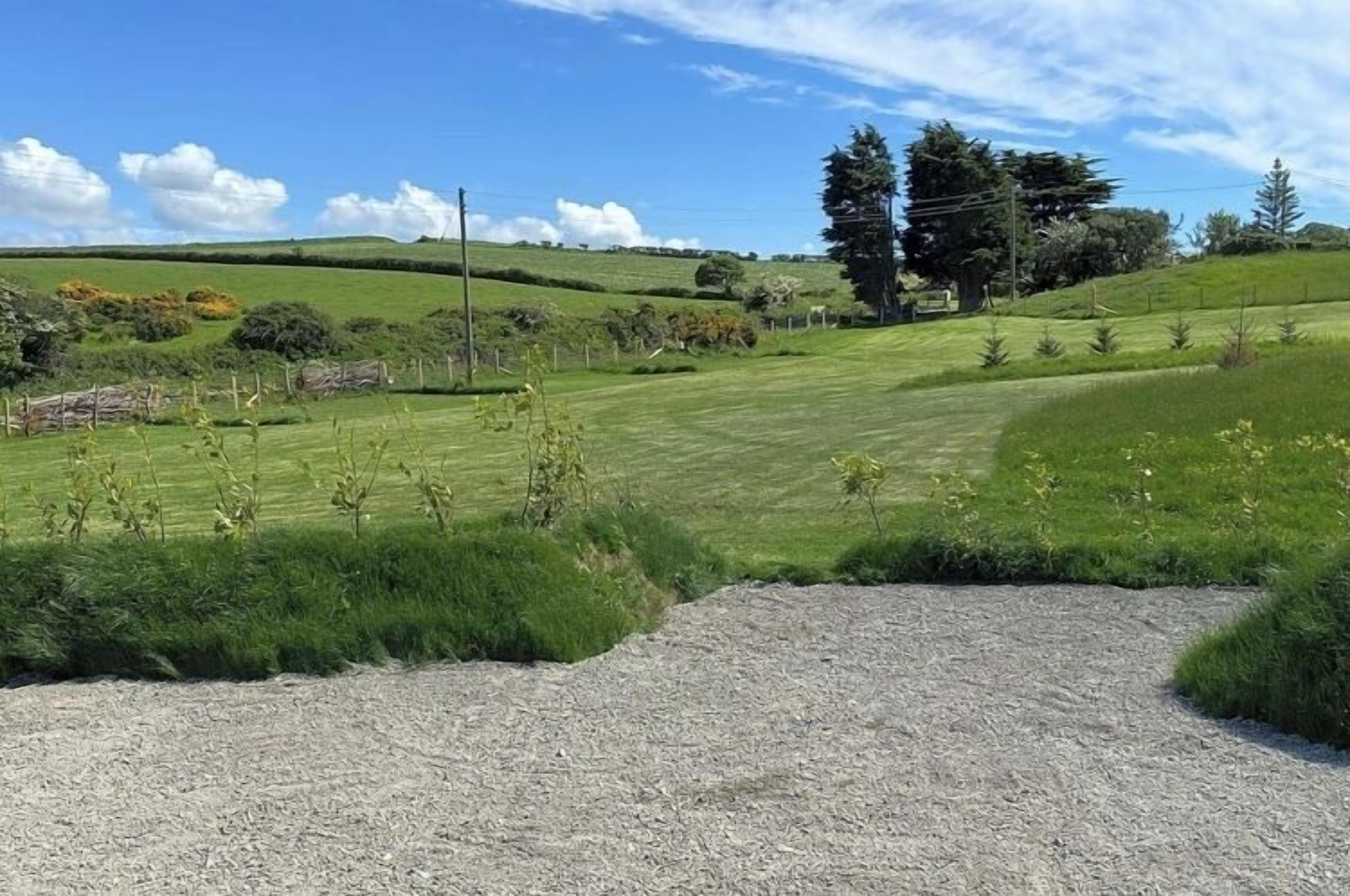 Paisaje rural con un camino de grava que atraviesa un campo de hierba, rodeado de árboles y colinas ondulantes.