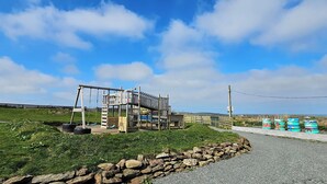 A playground with a wooden swing set and climbing structure, surrounded by a stone wall and a gravel path. - Cosy Annexe Sleeps 4 (Holyhead)