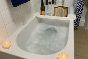 A bathtub with bubbles, a wine bottle, a glass, and a book on a shelf above it. - Cosy Annexe Sleeps 4 (Holyhead)