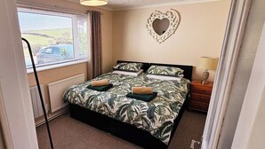 A bedroom with a bed, a heart-shaped mirror, a bedside table, and a window with blinds. - Cosy Annexe Sleeps 4 (Holyhead)
