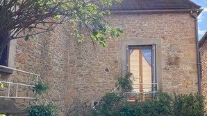 A stone building with a window and a door, surrounded by greenery and a paved path.
