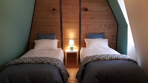 A room with two beds, each with a pillow and a bedside table with a lamp. The room has wooden paneling on the walls and a sloped ceiling.