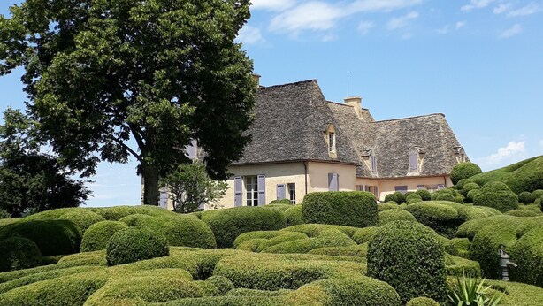 A landscaped garden with neatly trimmed hedges and a large tree in the foreground. In the background, there is a building with a stone roof and multiple windows.