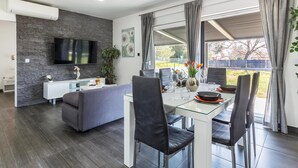 A modern living room with a grey sofa, a flat-screen TV mounted on a stone wall, a dining area with a glass table and black chairs, and a large window with curtains. - Beach front home in Peroj with house sea view (Peroj)